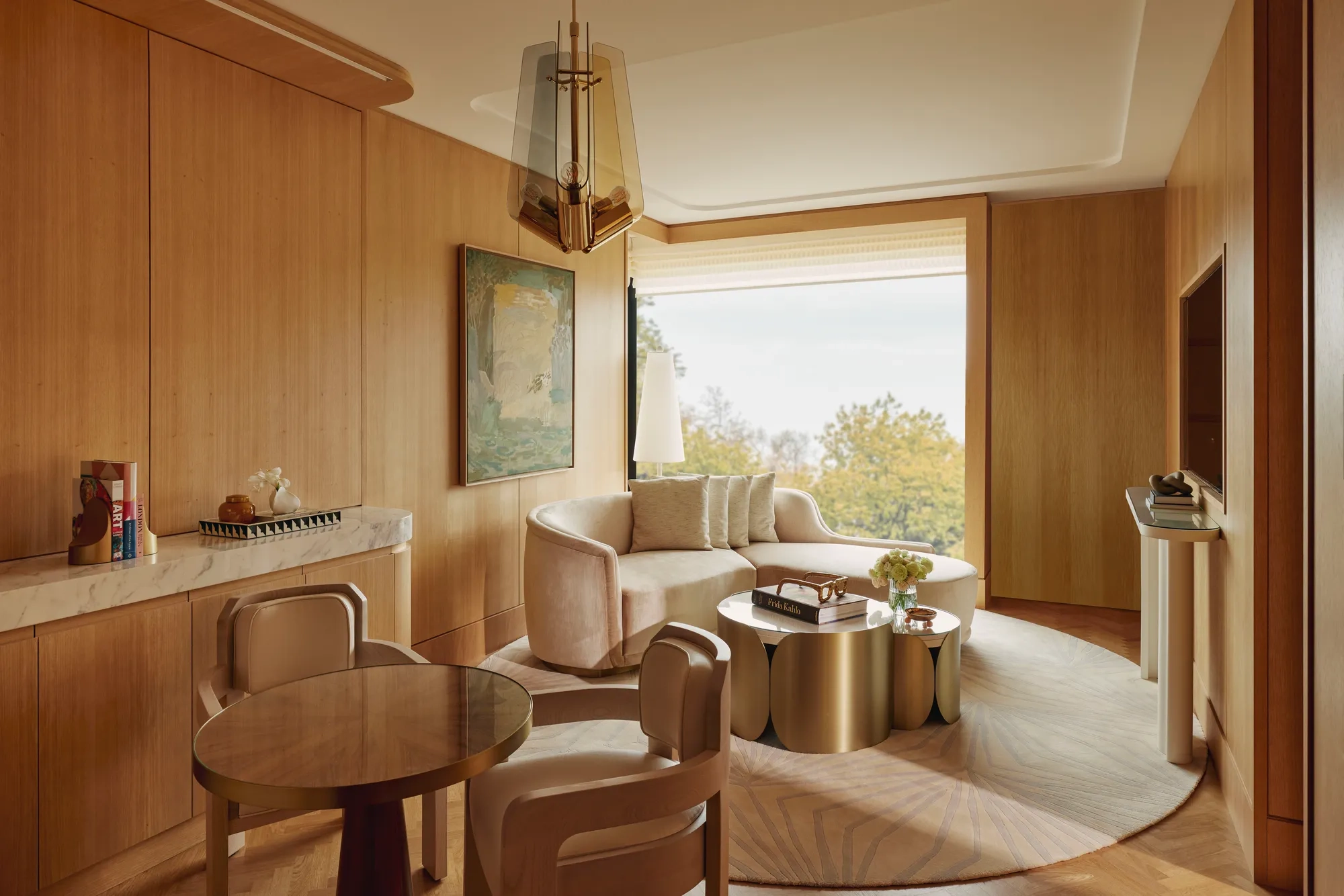 Warm wood-panelled suite living area with curved sofa, round dining table, brass coffee tables, and large window overlooking trees.