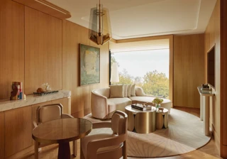 Warm wood-panelled suite living area with curved sofa, round dining table, brass coffee tables, and large window overlooking trees.