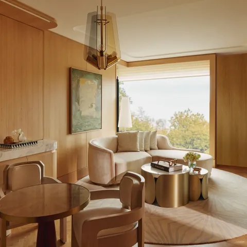 Warm wood-panelled suite living area with curved sofa, round dining table, brass coffee tables, and large window overlooking trees.