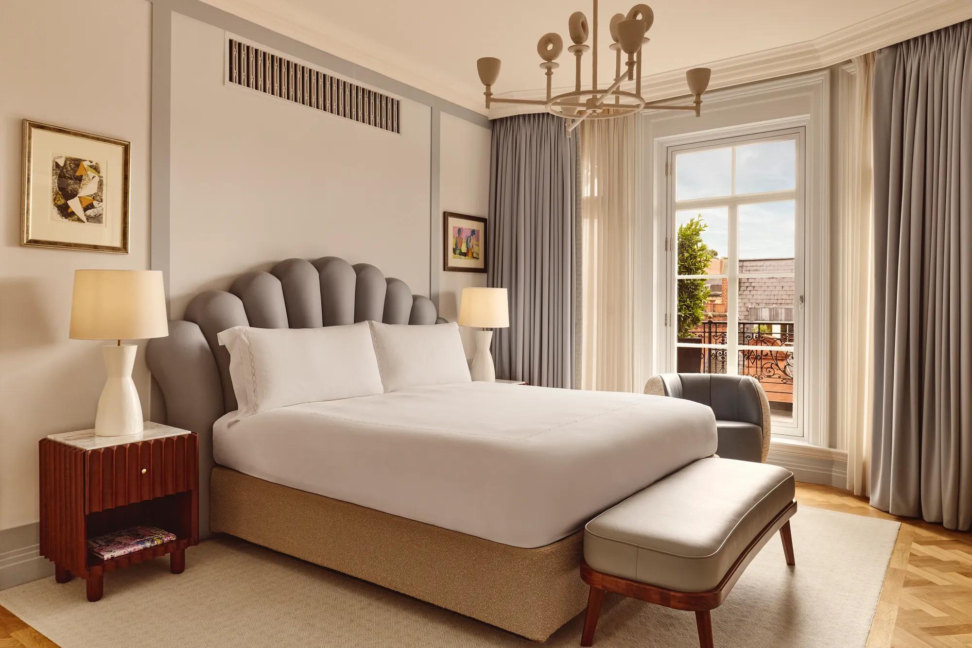 A bright bedroom featuring a large bed with a scalloped grey headboard, bedside tables with lamps, and floor-length curtains framing a window that opens to a terrace with red brick details.