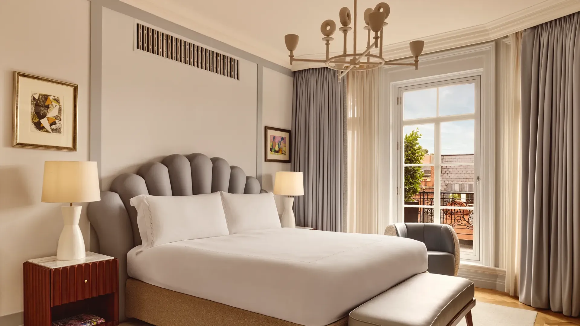 A bright bedroom featuring a large bed with a scalloped grey headboard, bedside tables with lamps, and floor-length curtains framing a window that opens to a terrace with red brick details.