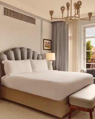 A bright bedroom featuring a large bed with a scalloped grey headboard, bedside tables with lamps, and floor-length curtains framing a window that opens to a terrace with red brick details.