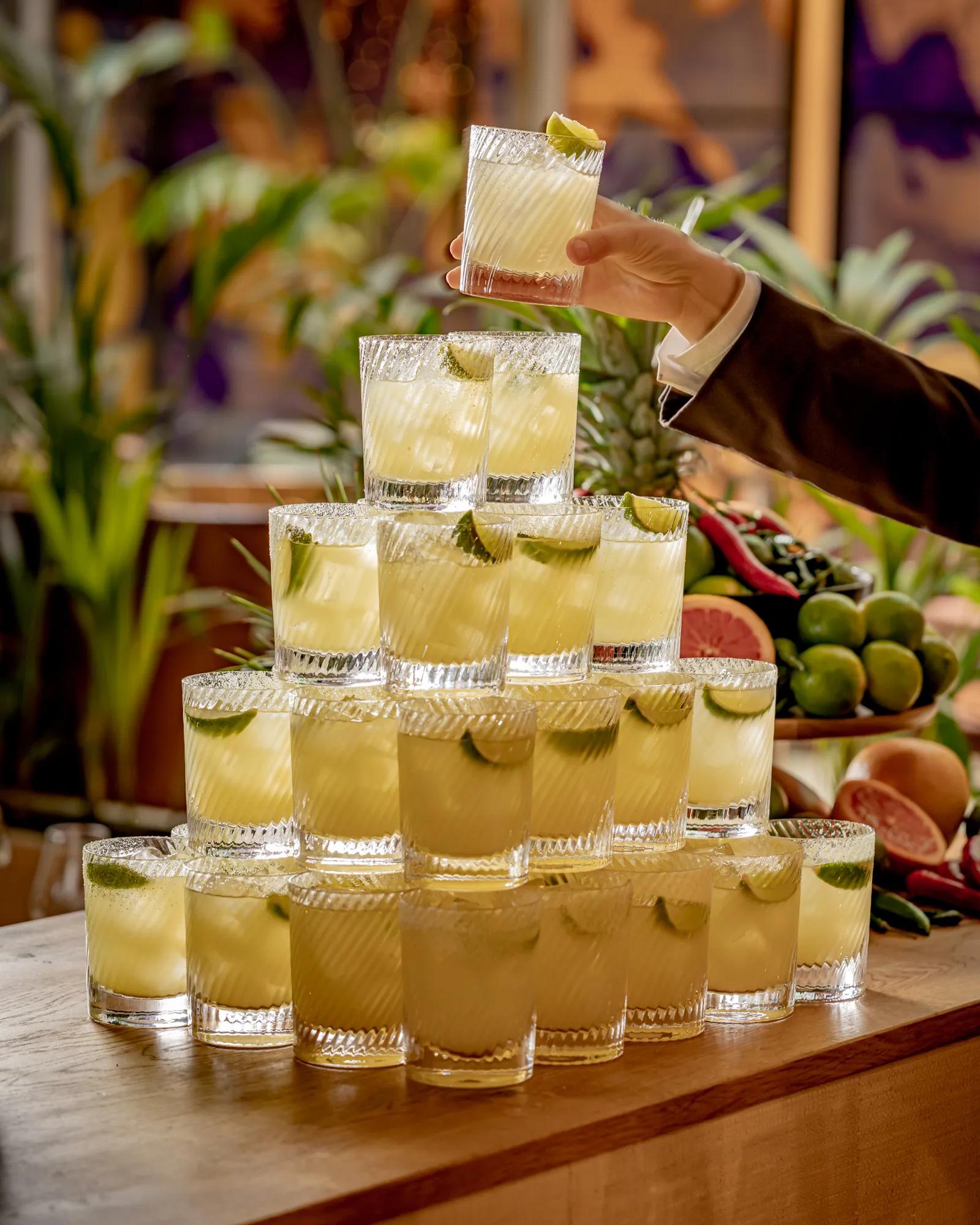 Pyramid of lime-garnished cocktails on a wooden bar, with a hand lifting a drink above the display
