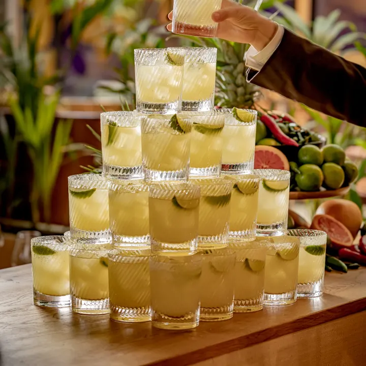 Pyramid of lime-garnished cocktails on a wooden bar, with a hand lifting a drink above the display