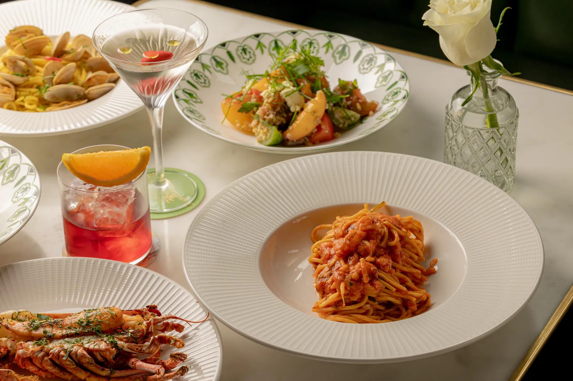 Tomato seafood pasta served on a white plate, surrounded by lobster, cocktails, salad, clams and a white rose on an elegant dining table.