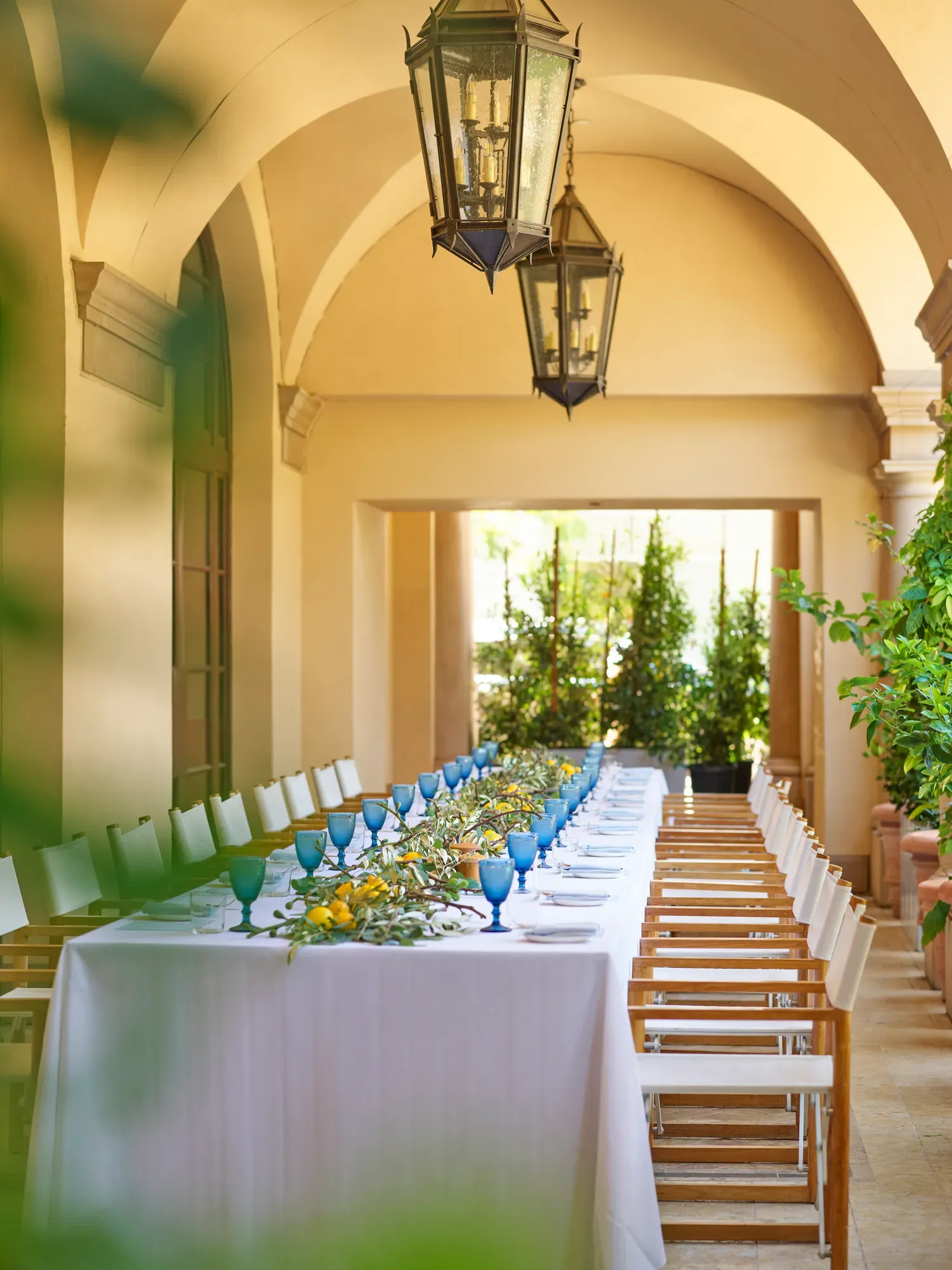 Portrait view down the Terrace Patio’s arched colonnade; lanterns above a long table dressed with blue goblets and lemon-eucalyptus garland.