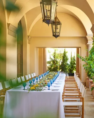 Portrait view down the Terrace Patio’s arched colonnade; lanterns above a long table dressed with blue goblets and lemon-eucalyptus garland.