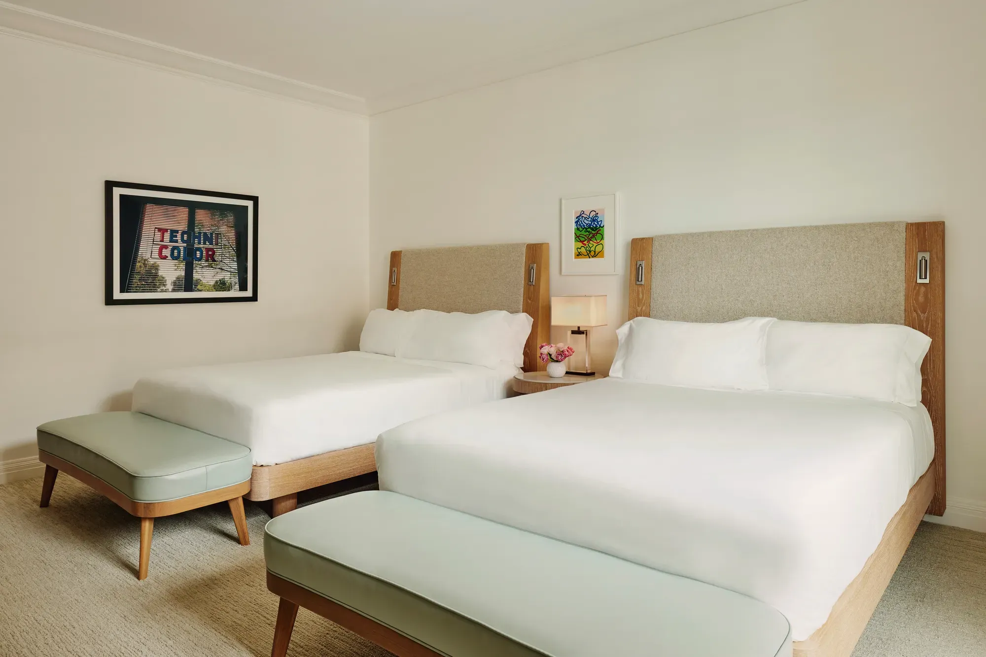 Two-queen room from opposite angle showing benches, central nightstand with lamp and flowers, and framed “Technicolor” artwork.