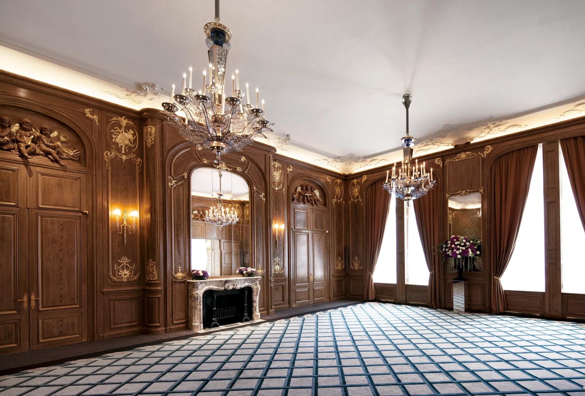 Ornate ballroom at Claridge’s with carved wood panelling, gold detailing, grand chandeliers, tall draped windows, and a decorative marble fireplace with a large mirror above.
