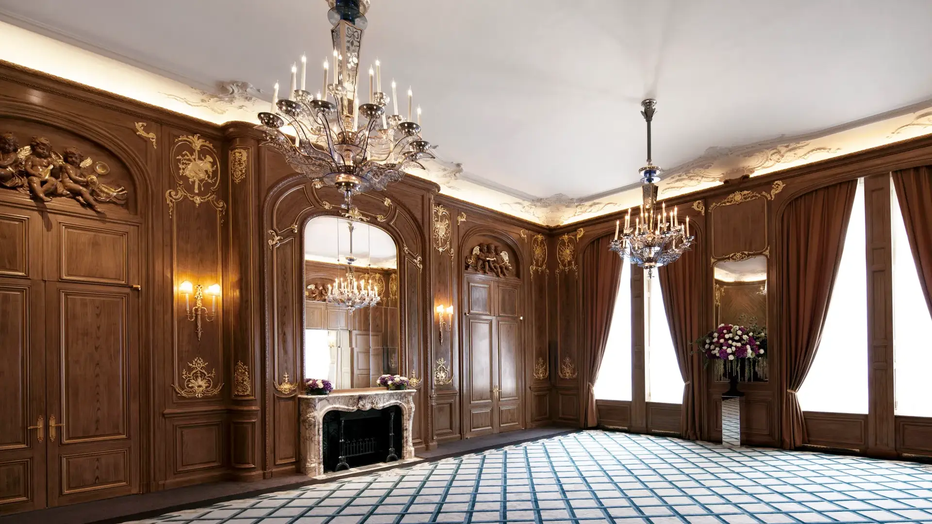 Ornate ballroom at Claridge’s with carved wood panelling, gold detailing, grand chandeliers, tall draped windows, and a decorative marble fireplace with a large mirror above.