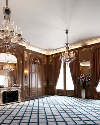 Ornate ballroom at Claridge’s with carved wood panelling, gold detailing, grand chandeliers, tall draped windows, and a decorative marble fireplace with a large mirror above.