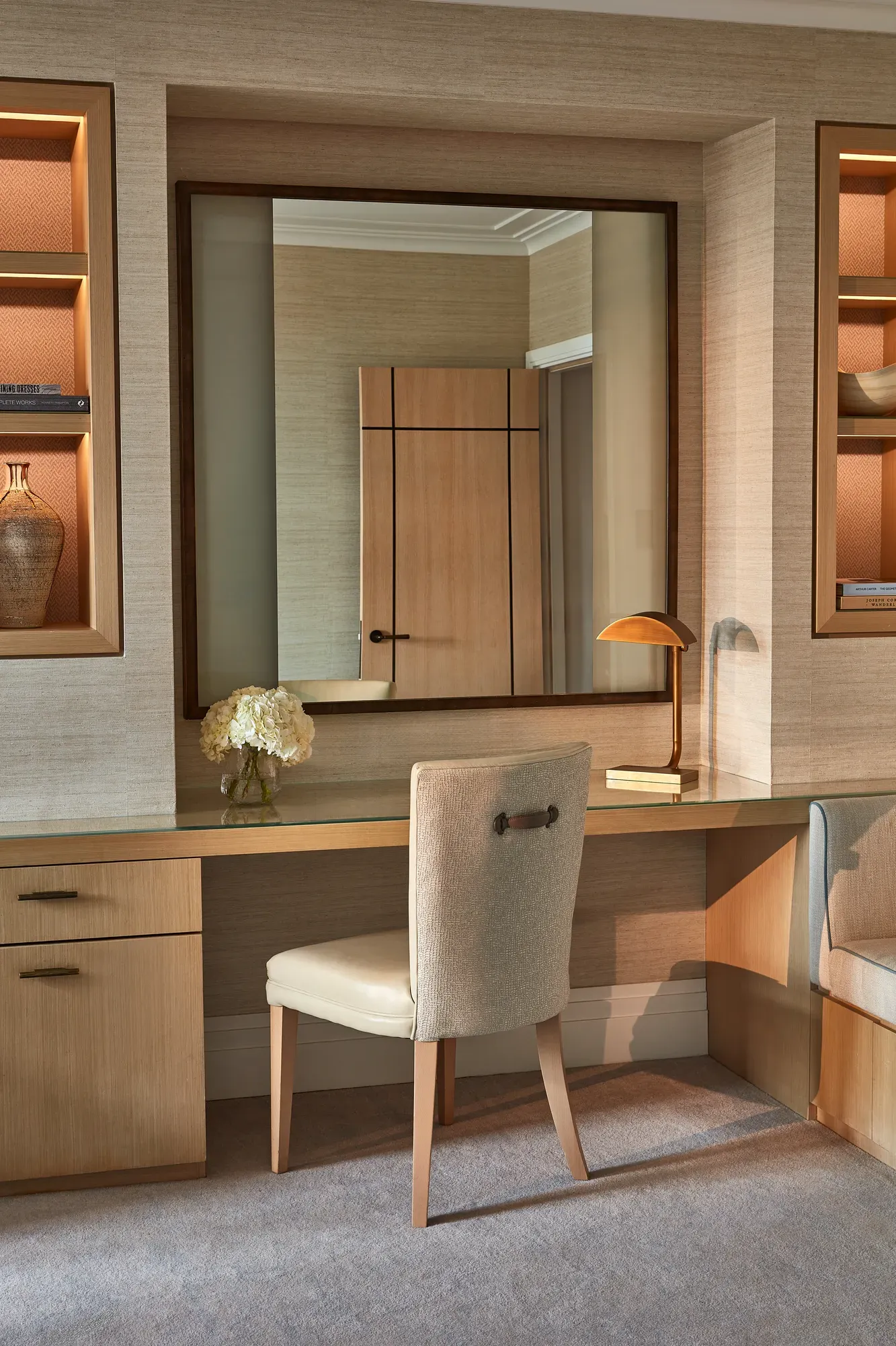 Built-in desk with upholstered chair, large mirror, shelves, and soft lighting in neutral tones.
