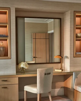 Built-in desk with upholstered chair, large mirror, shelves, and soft lighting in neutral tones.