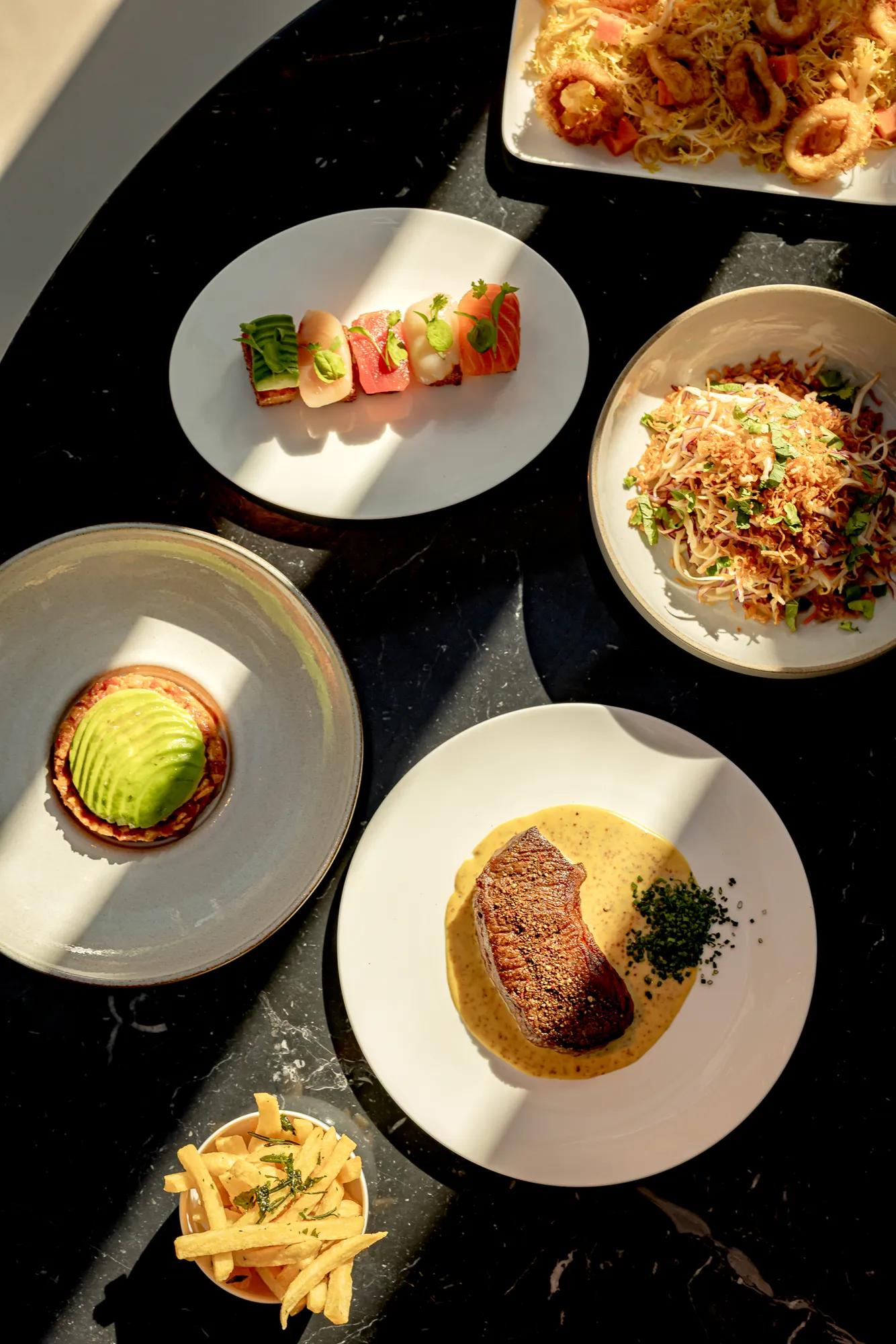 Overhead view of assorted restaurant dishes, including steak, sushi, noodles, avocado tart and fries, arranged on a dark marble table.