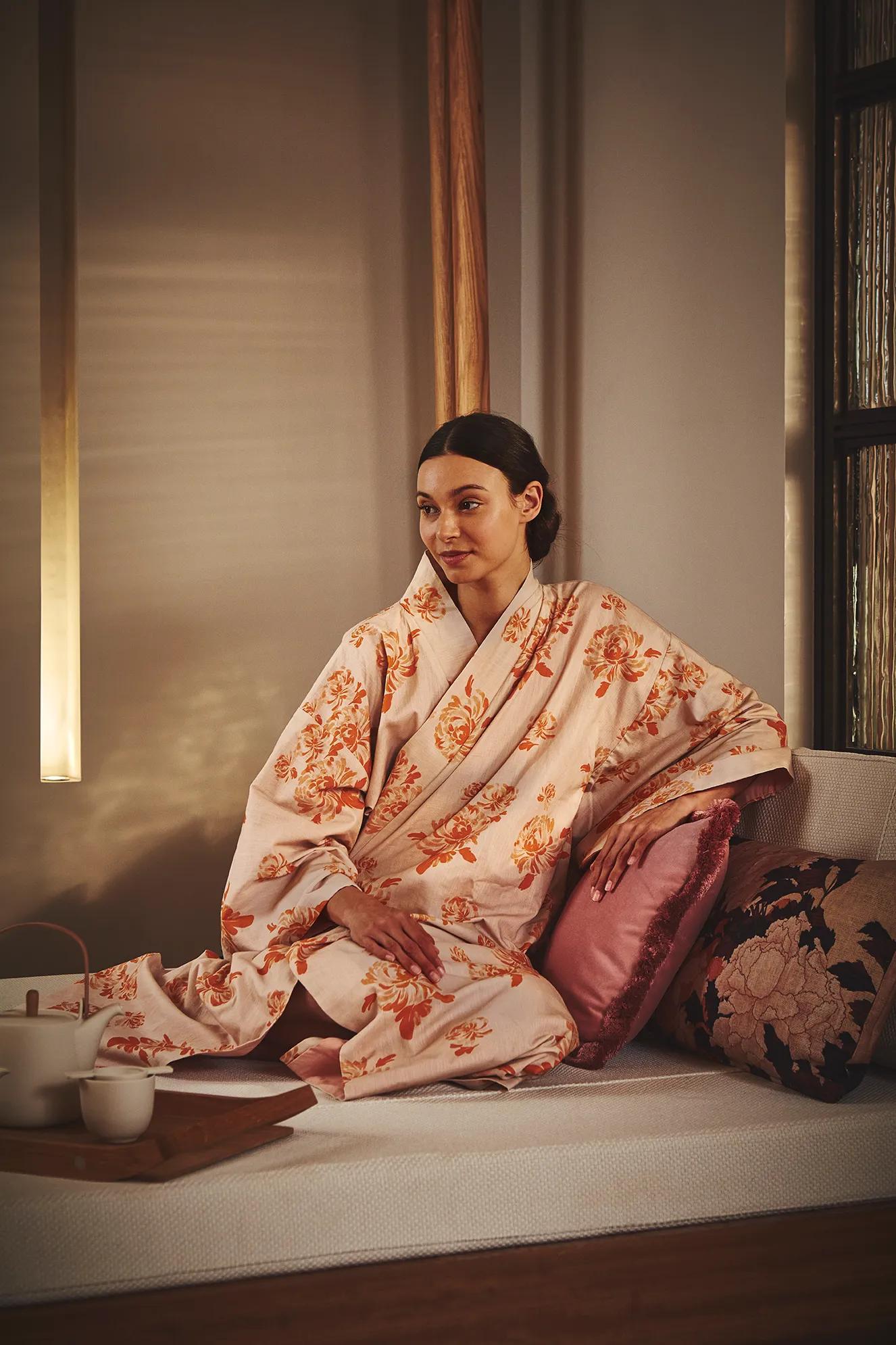 A woman wearing a floral kimono sits peacefully on a cushioned daybed with decorative pillows and a tea set beside her at The Maybourne Riviera spa.