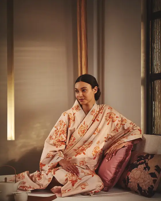 A woman wearing a floral kimono sits peacefully on a cushioned daybed with decorative pillows and a tea set beside her at The Maybourne Riviera spa.