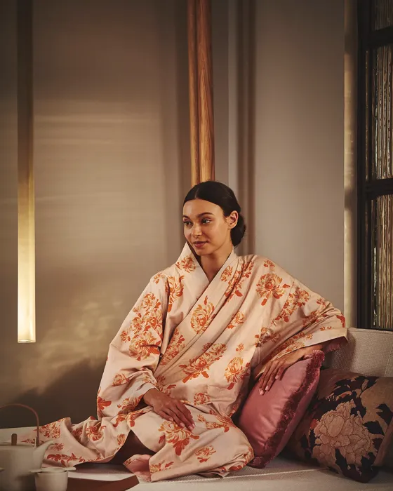 A woman wearing a floral kimono sits peacefully on a cushioned daybed with decorative pillows and a tea set beside her at The Maybourne Riviera spa.