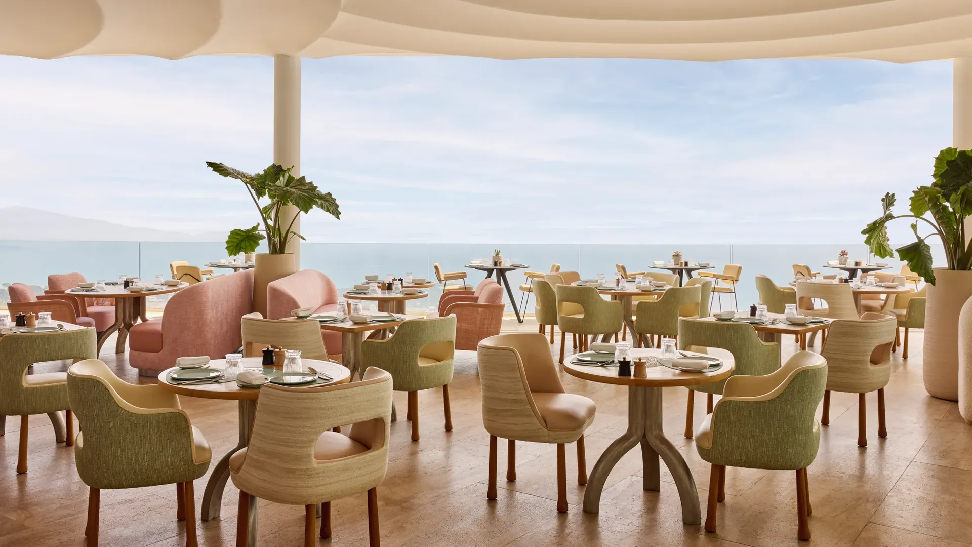 Restaurant élégant avec des chaises aux tons pastel et des tables rondes dressées avec raffinement, offrant une vue sur la mer à travers de grandes baies vitrées.