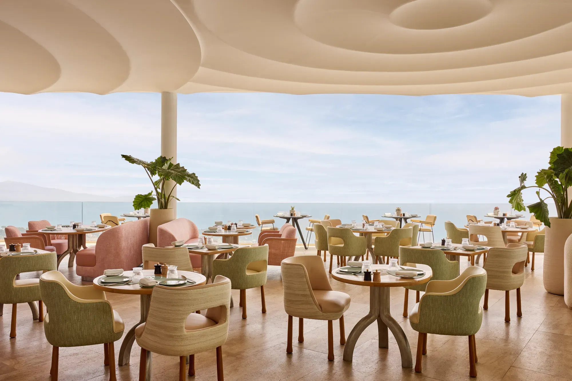 Restaurant élégant avec des chaises aux tons pastel et des tables rondes dressées avec raffinement, offrant une vue sur la mer à travers de grandes baies vitrées.