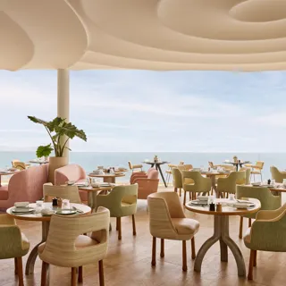 Restaurant élégant avec des chaises aux tons pastel et des tables rondes dressées avec raffinement, offrant une vue sur la mer à travers de grandes baies vitrées.