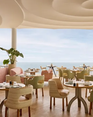 Restaurant élégant avec des chaises aux tons pastel et des tables rondes dressées avec raffinement, offrant une vue sur la mer à travers de grandes baies vitrées.