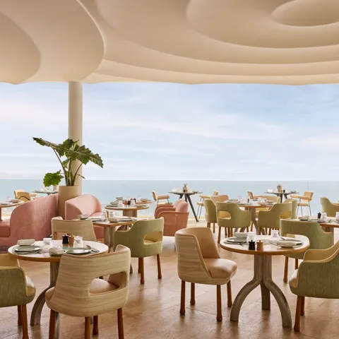 Elegant restaurant with pastel-colored chairs and round tables set with fine tableware, overlooking the sea through floor-to-ceiling windows.