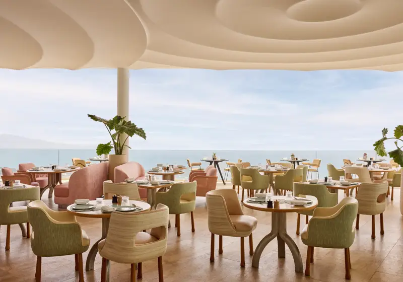 Restaurant élégant avec des chaises aux tons pastel et des tables rondes dressées avec raffinement, offrant une vue sur la mer à travers de grandes baies vitrées.