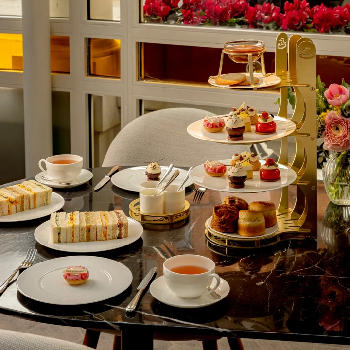 Afternoon tea set with tiered pastries, finger sandwiches, and cups of tea on a marble table by a window with pink flowers.