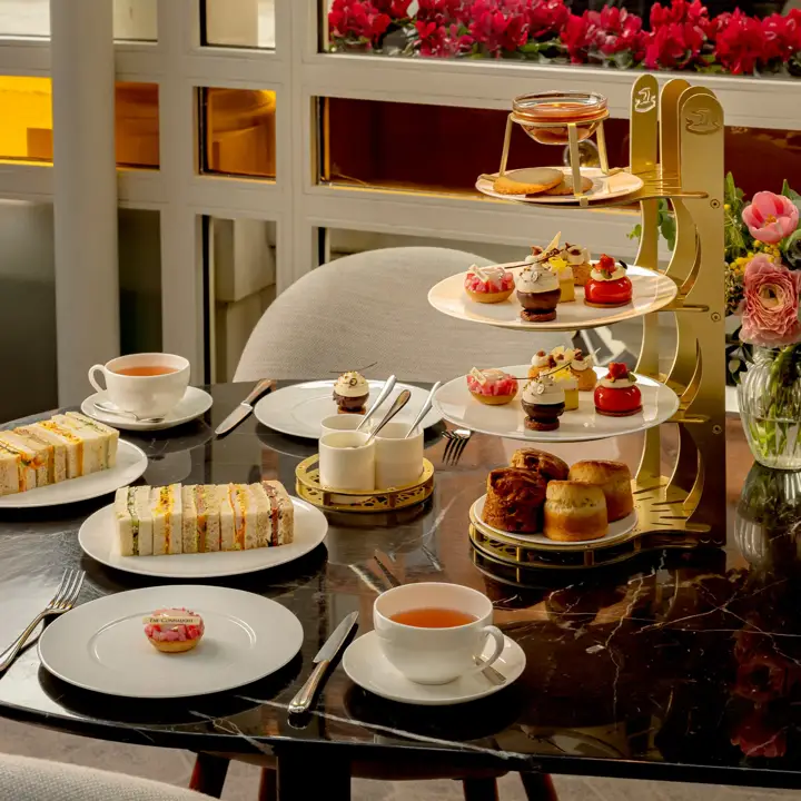 Afternoon tea set with tiered pastries, finger sandwiches, and cups of tea on a marble table by a window with pink flowers.