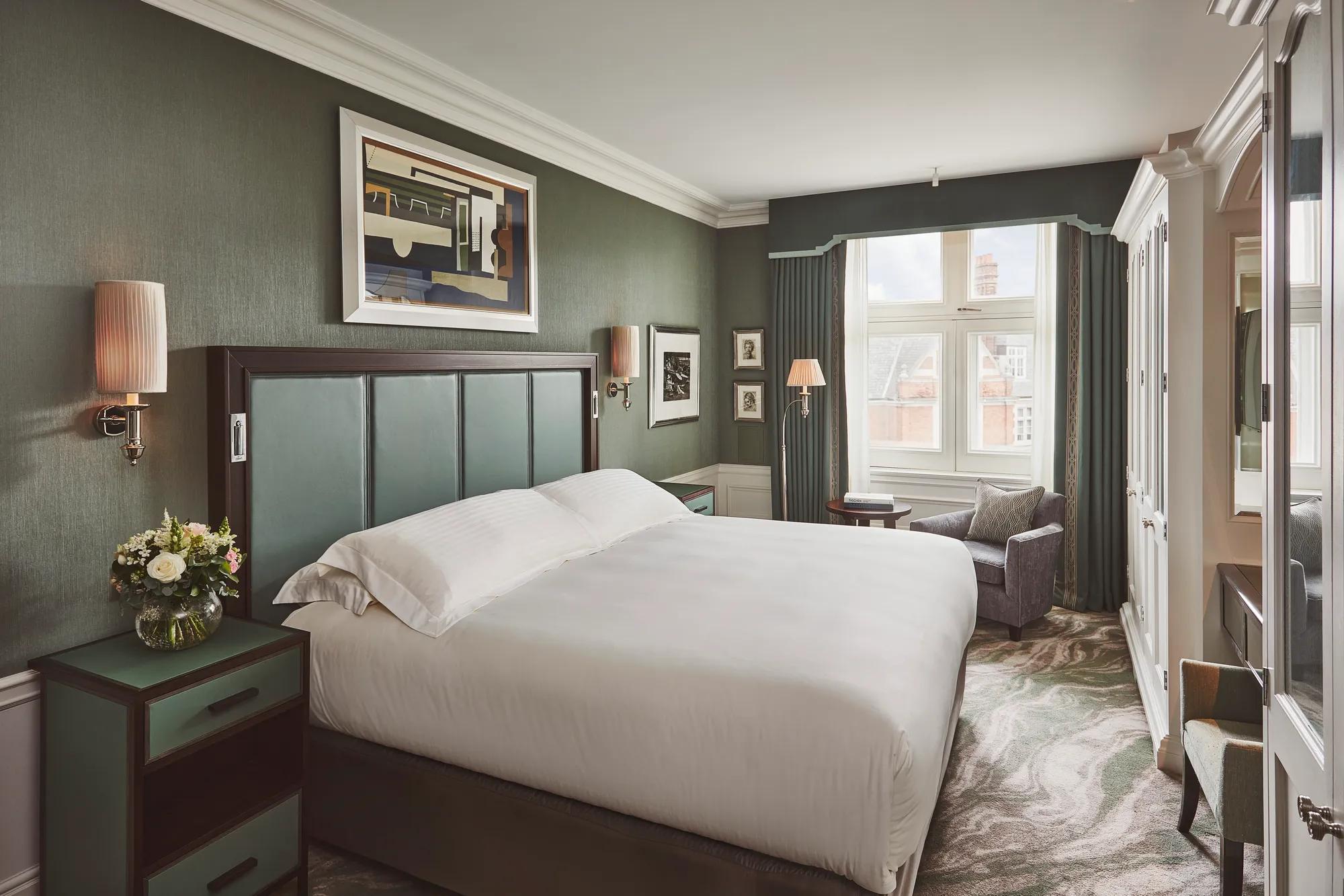 Bedroom with large upholstered bed, green walls, bedside flowers, and a window with armchair and table.