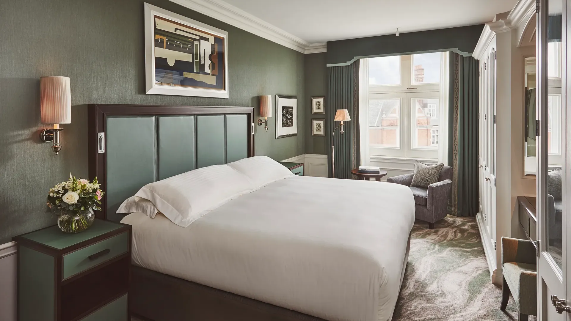 Bedroom with large upholstered bed, green walls, bedside flowers, and a window with armchair and table.