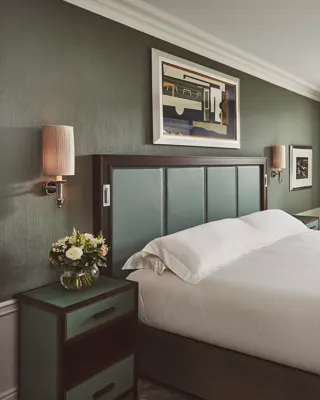 Bedroom with large upholstered bed, green walls, bedside flowers, and a window with armchair and table.