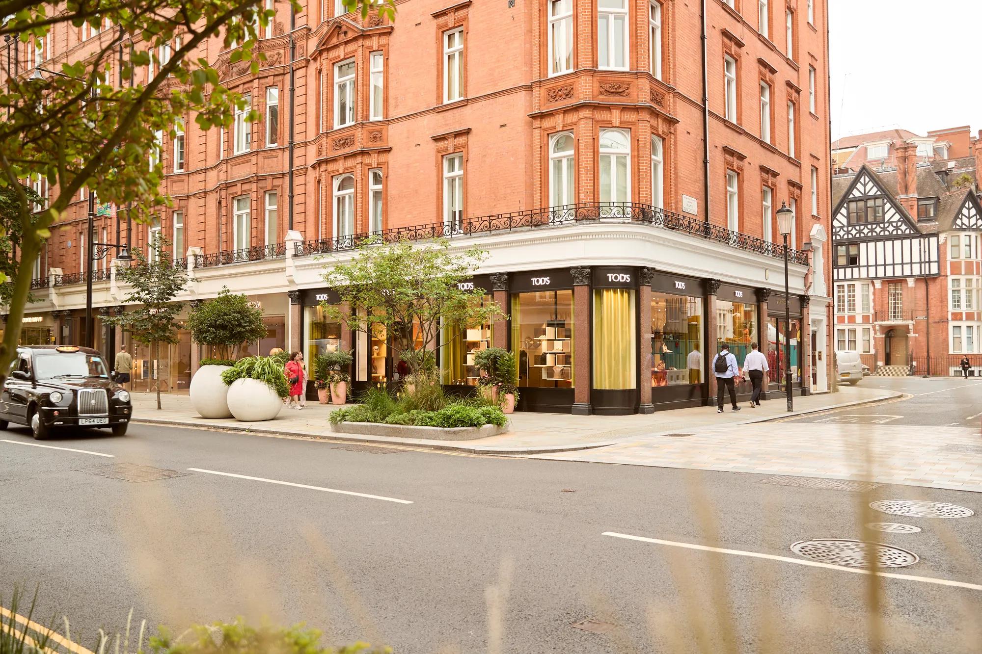 Corner boutique storefront in Mayfair, London, with elegant red-brick architecture, luxury retail displays, landscaped planters, and a classic black cab passing by.