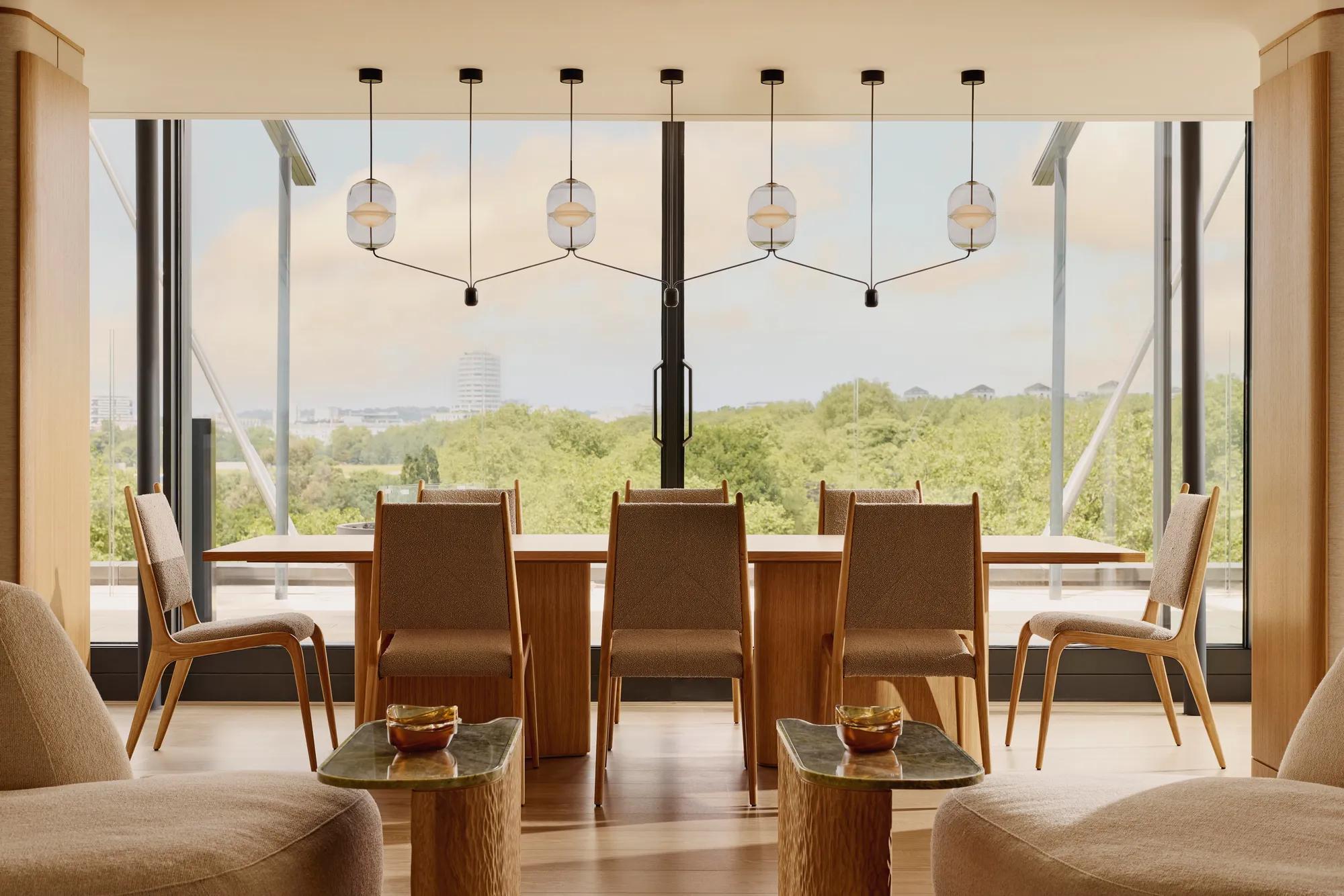 A modern dining area with a long wooden table and eight chairs, framed by pendant lights and full-height windows overlooking Hyde Park and the London skyline.