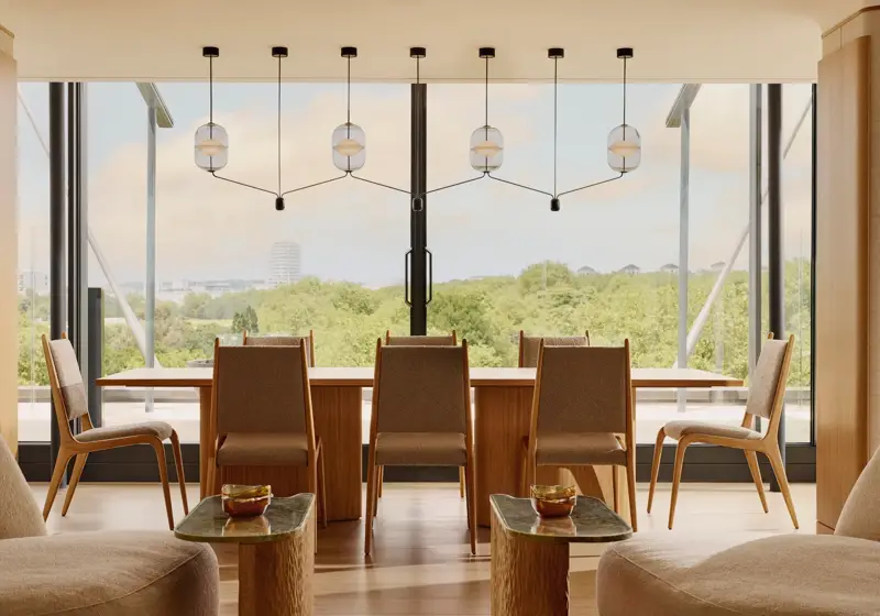A modern dining area with a long wooden table and eight chairs, framed by pendant lights and full-height windows overlooking Hyde Park and the London skyline.