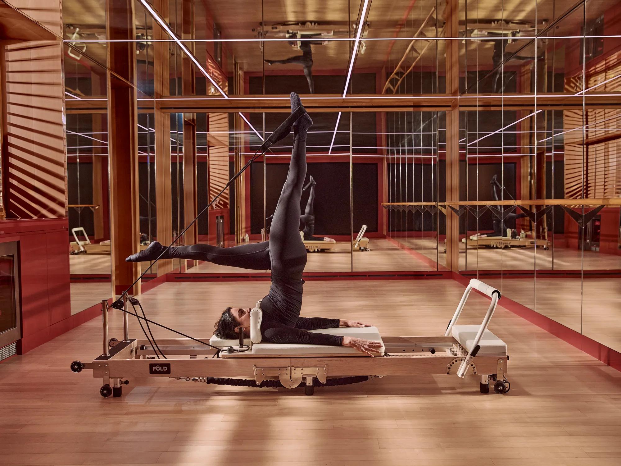 Person performing a Pilates reformer exercise in a mirrored studio, extending one leg overhead while using resistance straps on specialised equipment.