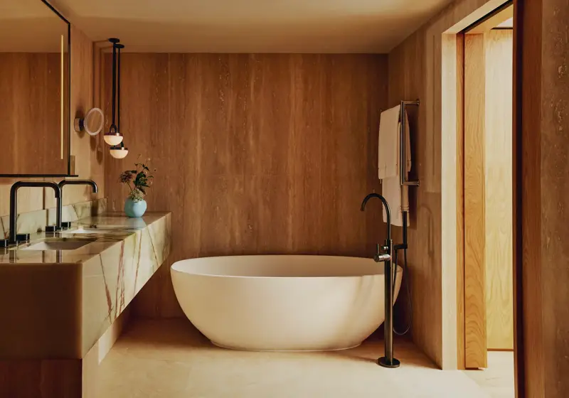 Warm-toned bathroom with a freestanding oval bathtub, marble vanity with twin basins, and black fittings against wood-panelled walls.