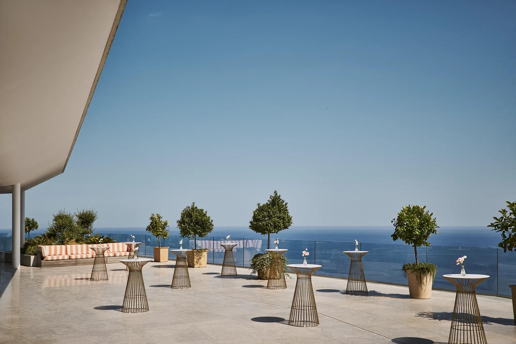 Grande terrasse avec tables hautes, arbres en pots et banquette rayée, offrant une vue sur la mer bleue sous un ciel clair.