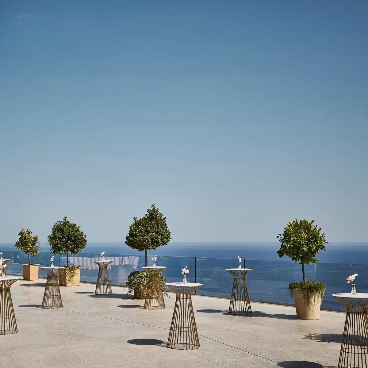 Spacious terrace with high cocktail tables, potted trees and striped seating area, overlooking a clear blue sea under a bright sky.