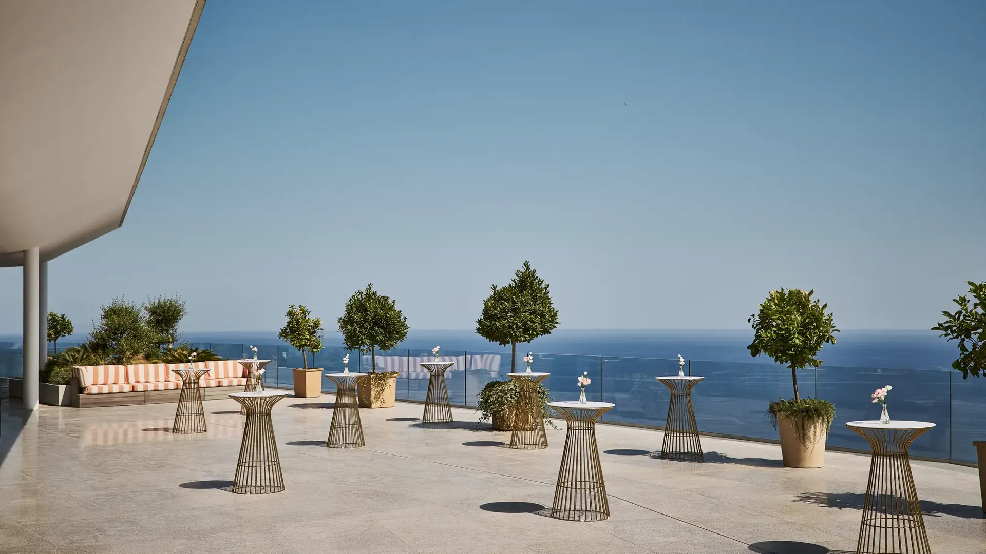 Grande terrasse avec tables hautes, arbres en pots et banquette rayée, offrant une vue sur la mer bleue sous un ciel clair.