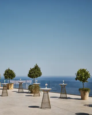 Grande terrasse avec tables hautes, arbres en pots et banquette rayée, offrant une vue sur la mer bleue sous un ciel clair.