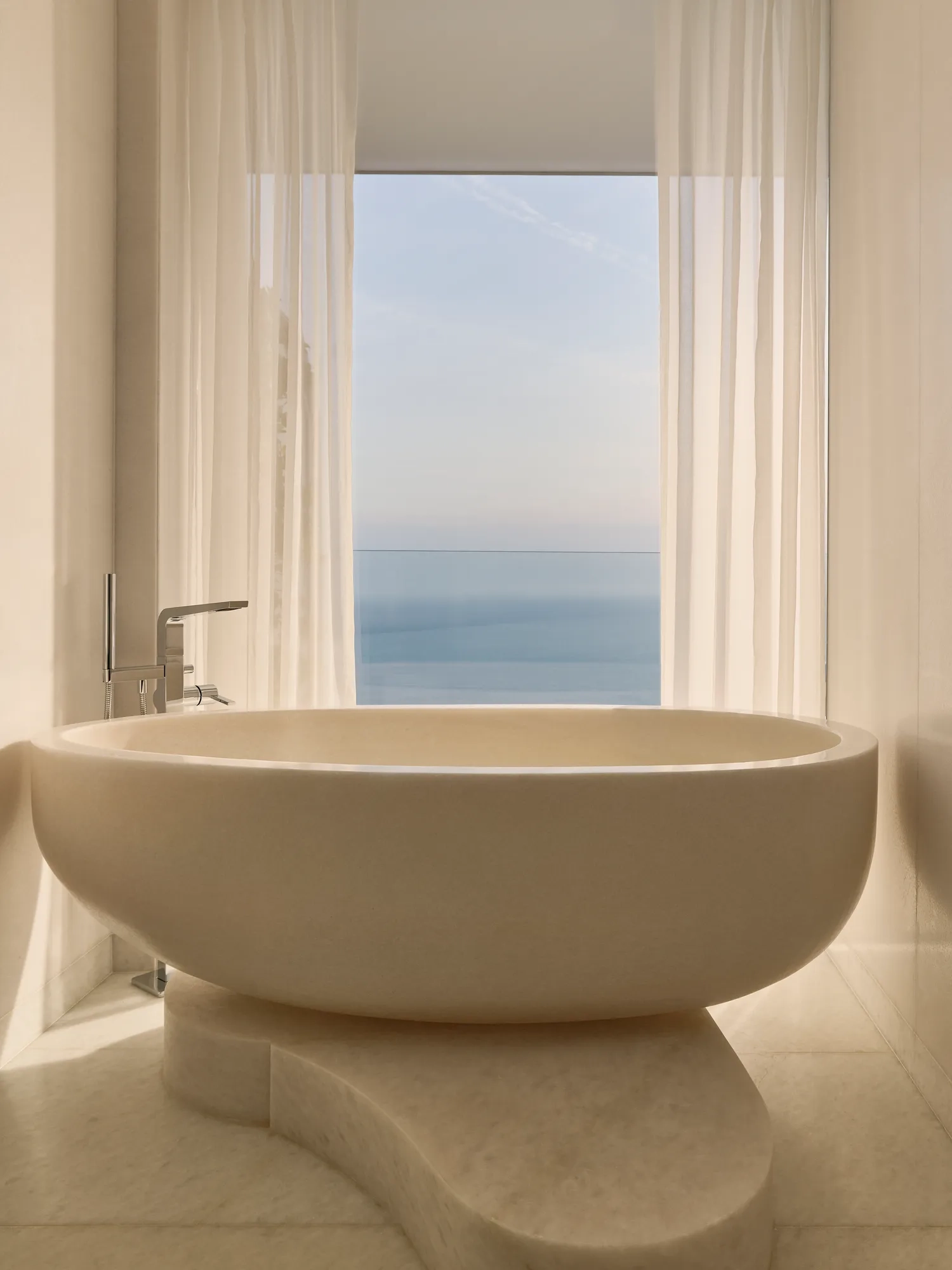 Salle de bain élégante avec une baignoire autoportante sculpturale posée sur un socle en pierre incurvé, encadrée de voilages et d’une fenêtre offrant une vue apaisante sur la mer.