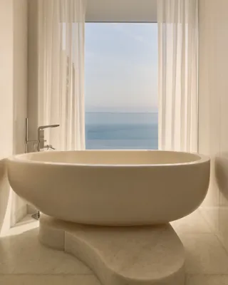Elegant bathroom with a sculptural freestanding bathtub positioned on a curved stone base, framed by sheer curtains and a window offering a serene sea view.