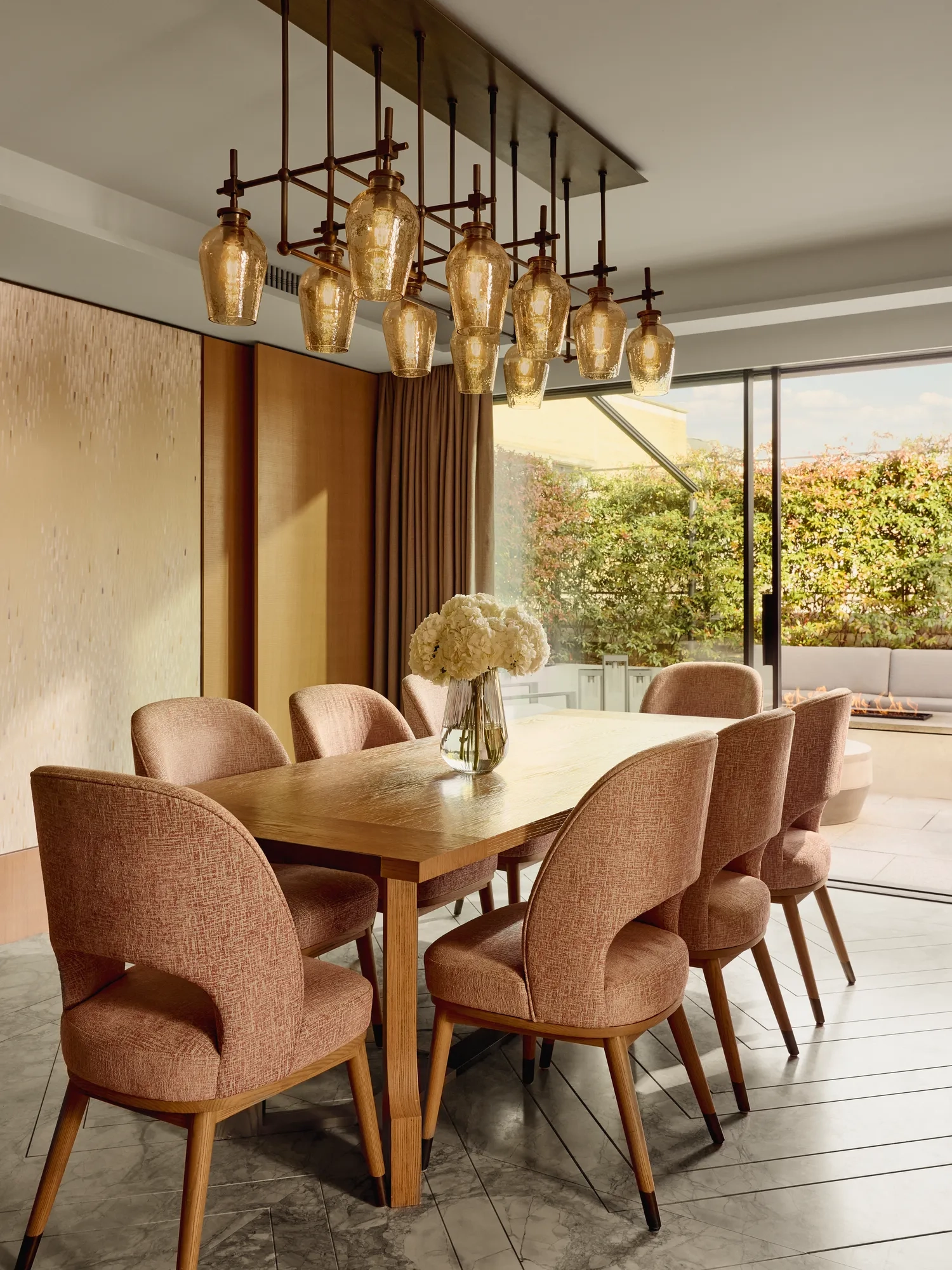 Dining room with wooden table, pink upholstered chairs, and modern glass pendant chandelier, opening to terrace