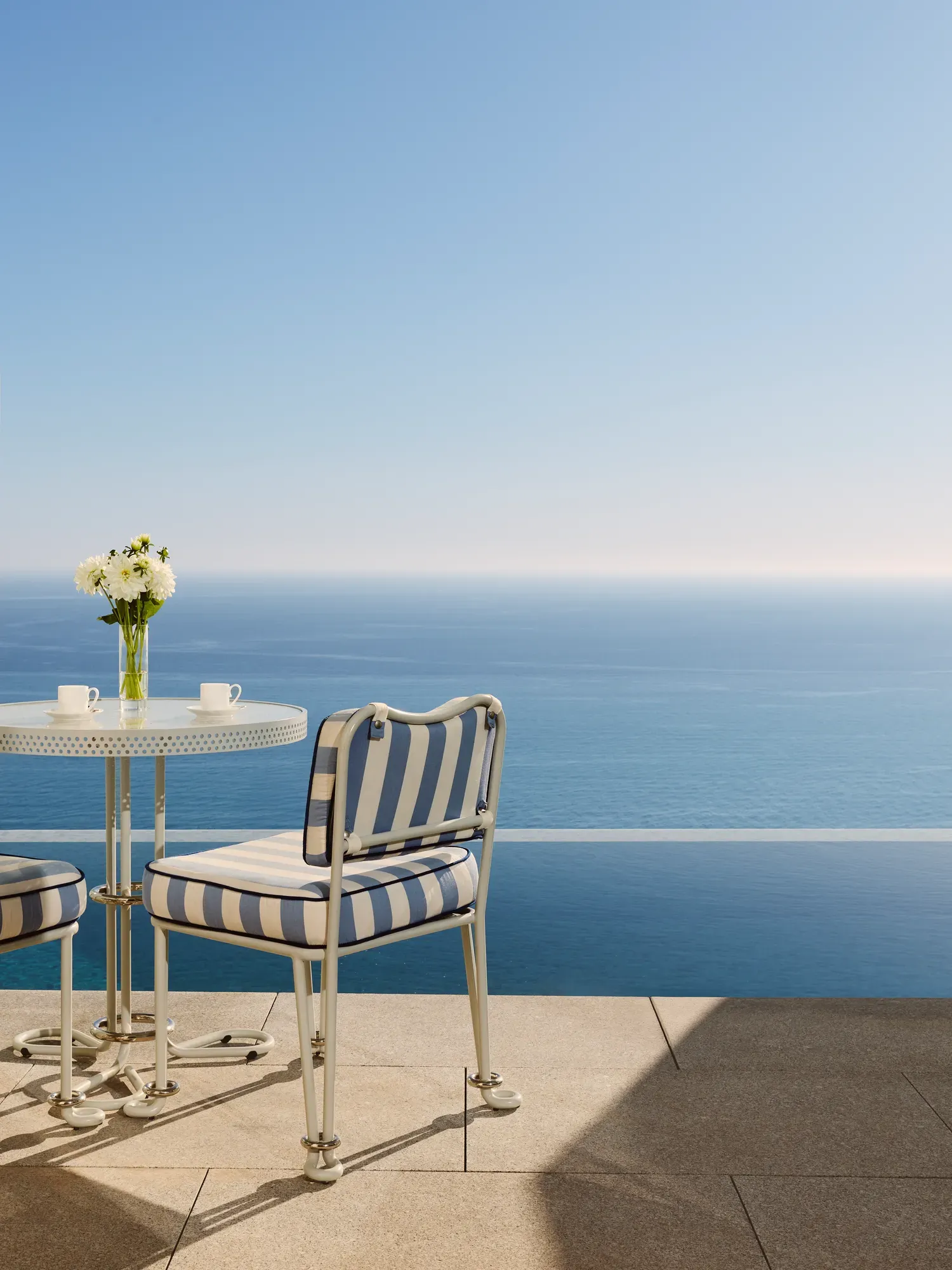 Terrace table with striped chairs and fresh flowers overlooks the calm blue sea under a clear sky.