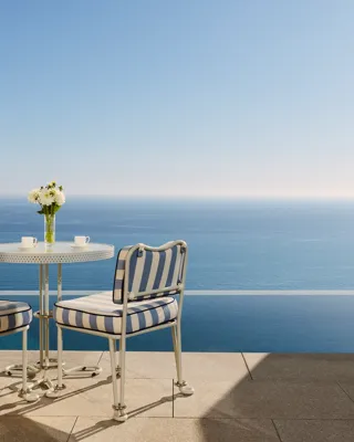 Table de terrasse avec chaises rayées et fleurs fraîches donnant sur la mer bleue calme sous un ciel dégagé.