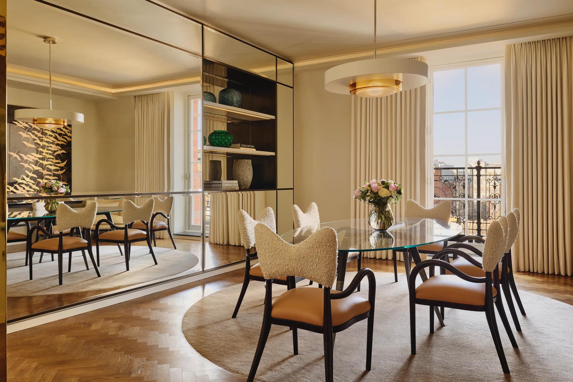 Sophisticated dining room with a glass-topped oval table surrounded by cream and tan chairs, a mirrored wall reflecting natural light, and a modern chandelier hanging above a neutral rug.