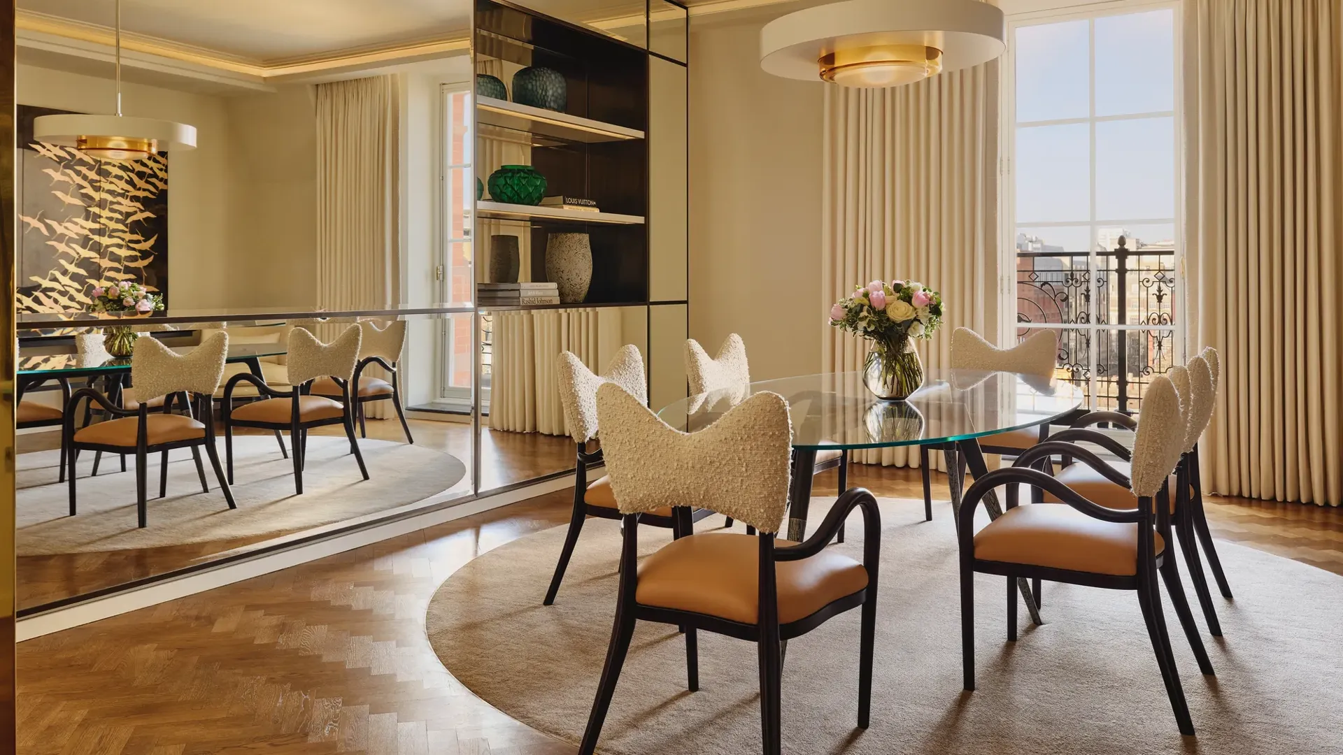 Sophisticated dining room with a glass-topped oval table surrounded by cream and tan chairs, a mirrored wall reflecting natural light, and a modern chandelier hanging above a neutral rug.