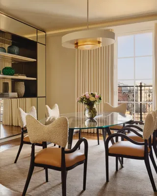 Sophisticated dining room with a glass-topped oval table surrounded by cream and tan chairs, a mirrored wall reflecting natural light, and a modern chandelier hanging above a neutral rug.