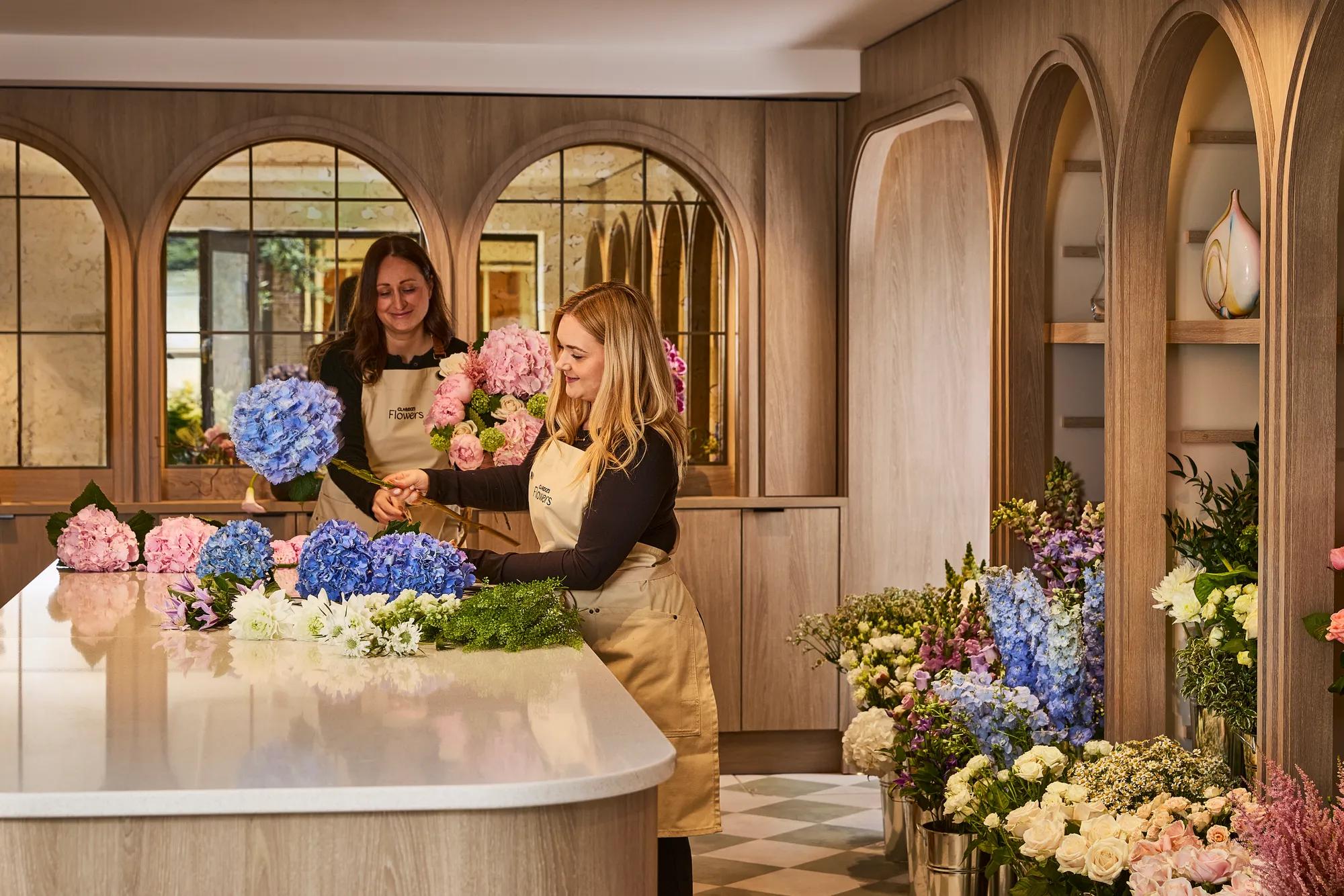 **Alt text (WCAG 2.2 AA, under 125 characters):**

Two florists arrange fresh bouquets on a central worktable, surrounded by seasonal flowers in a warm, wood-panelled boutique.
